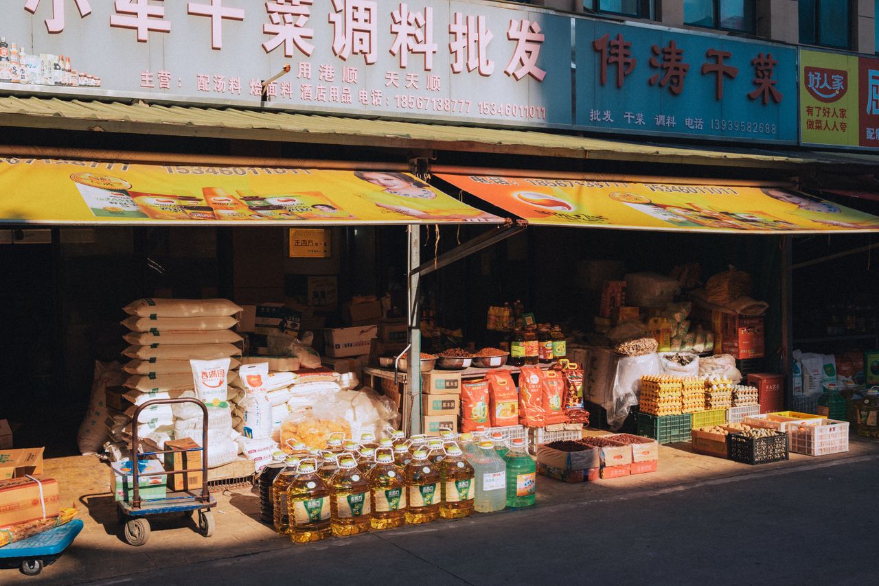Variety of products displayed in a Chinese market stall under bright sunlight, featuring groceries and packaging.